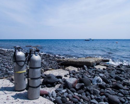 Sidemount cylinders stand on a pebbly beach, pre-prepared by a diver that is qualified in solo diving to go for a dive on their own